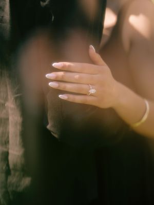 Close-up of a womans hand wearing a delicate diamond ring with long nude nails, resting on a dark surface.