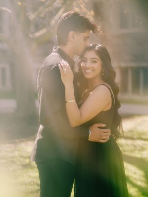 A couple embraces outdoors in golden sunlight, the woman smiling in a black dress while the man holds her close.