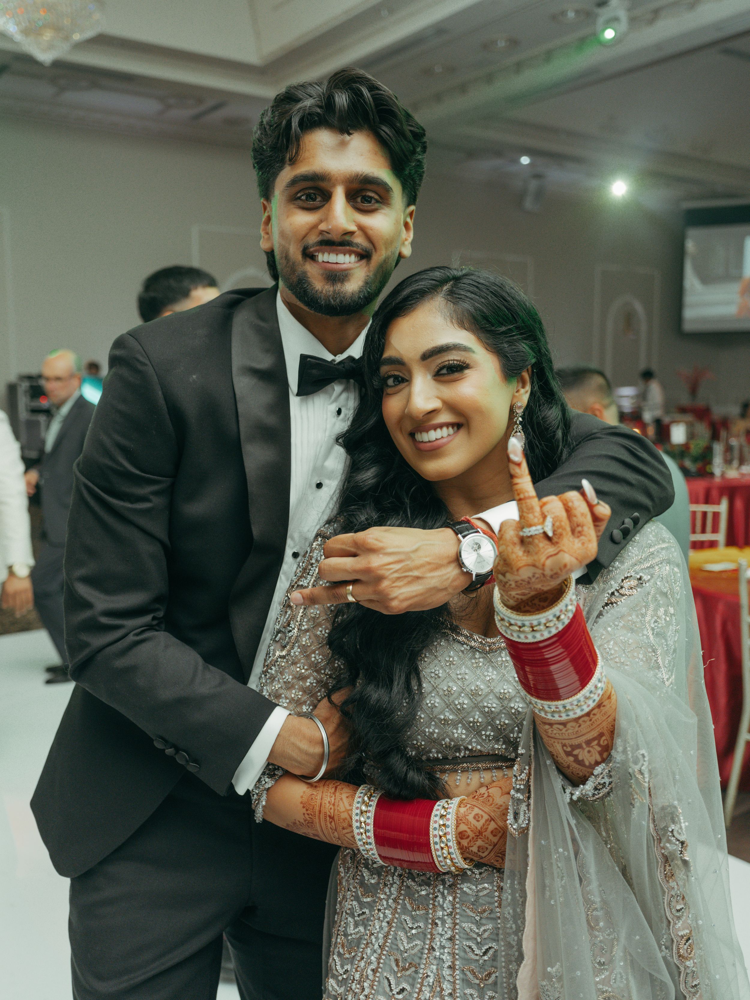 Couple in wedding attire posing at reception, groom in tuxedo embracing bride in embroidered lehenga with mehndi.