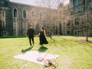Couple in black outfits holding hands on grass near a stone university building with a picnic blanket nearby.