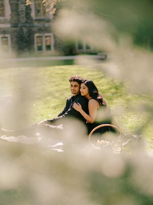 Couple sitting together on grass lawn embracing during outdoor engagement photo session near historic stone building.