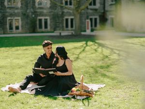 Couple in black outfits enjoying a romantic picnic on green grass outside a stone building.