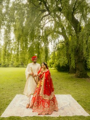Indian bride and groom in traditional red lehenga and white sherwani standing under a willow tree on a rug.