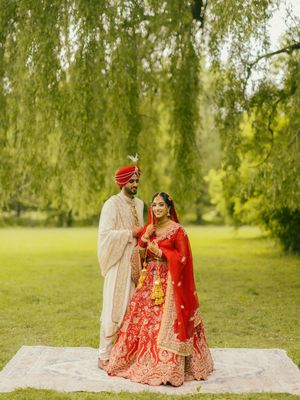 Indian bride in red lehenga and groom in cream sherwani pose under willow tree during outdoor wedding.