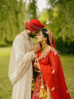 South Asian bride and groom in traditional red and gold wedding attire share a romantic moment outdoors.
