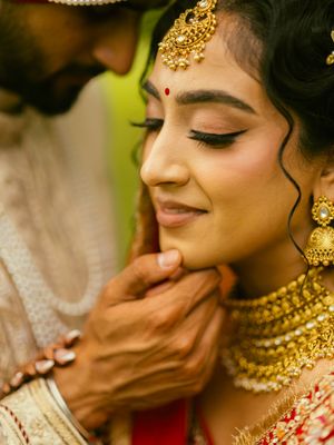 Indian bride in gold jewelry and red bindi smiles as groom tenderly holds her face during wedding ceremony.