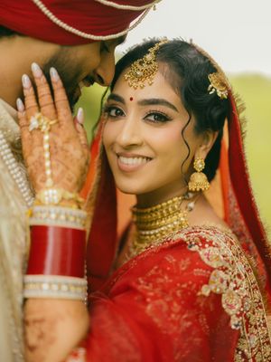 Indian bride in red and gold lehenga with traditional jewelry smiles beside groom during wedding ceremony.