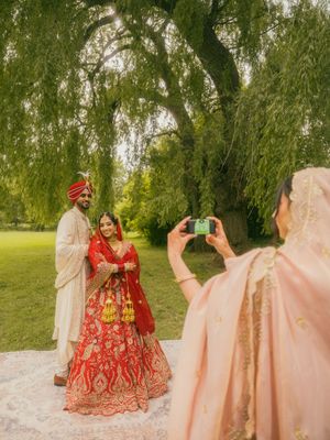 Guest photographs Indian wedding couple in traditional attire under a willow tree outdoors.