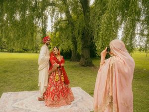 A woman photographs a Sikh bride in red lehenga and groom in cream sherwani standing on a blanket under a willow tree.