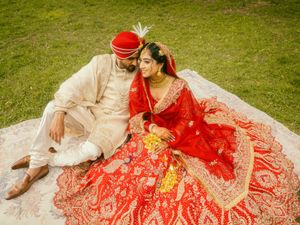 Indian bride in red lehenga and groom in cream sherwani sitting together on white cloth during wedding ceremony.