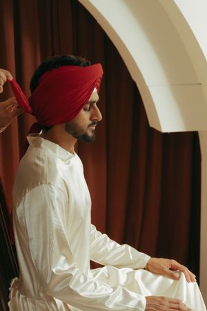 A young South Asian man in white kurta sits as someone ties a red turban around his head.