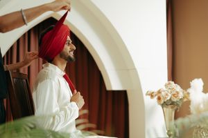 A Sikh groom smiles as someone ties his red turban, wearing a white sherwani in an elegant indoor setting.