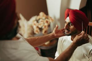 Two Sikh men in red turbans share a warm moment, one gently touching the other's face indoors.