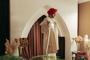 A person in a white robe and red hat poses elegantly under an arch, surrounded by floral arrangements.