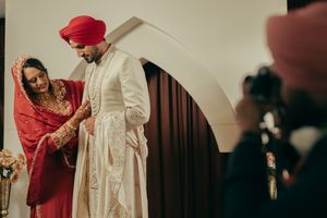 Sikh bride in red dupatta helps groom in white sherwani dress during traditional wedding ceremony.