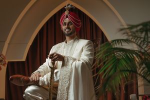 A groom in traditional South Asian white sherwani and red turban sits elegantly in an ornate chair during wedding ceremony.