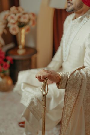 Groom in white embroidered sherwani holding a decorative sword, seated beside floral arrangements.