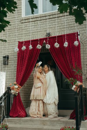 Two women in traditional South Asian attire pose for a selfie under elegant red velvet curtains at a wedding ceremony entrance.
