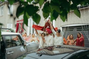 Indian groom in red turban shielded by red cloth during traditional wedding ceremony outdoors near decorated home.