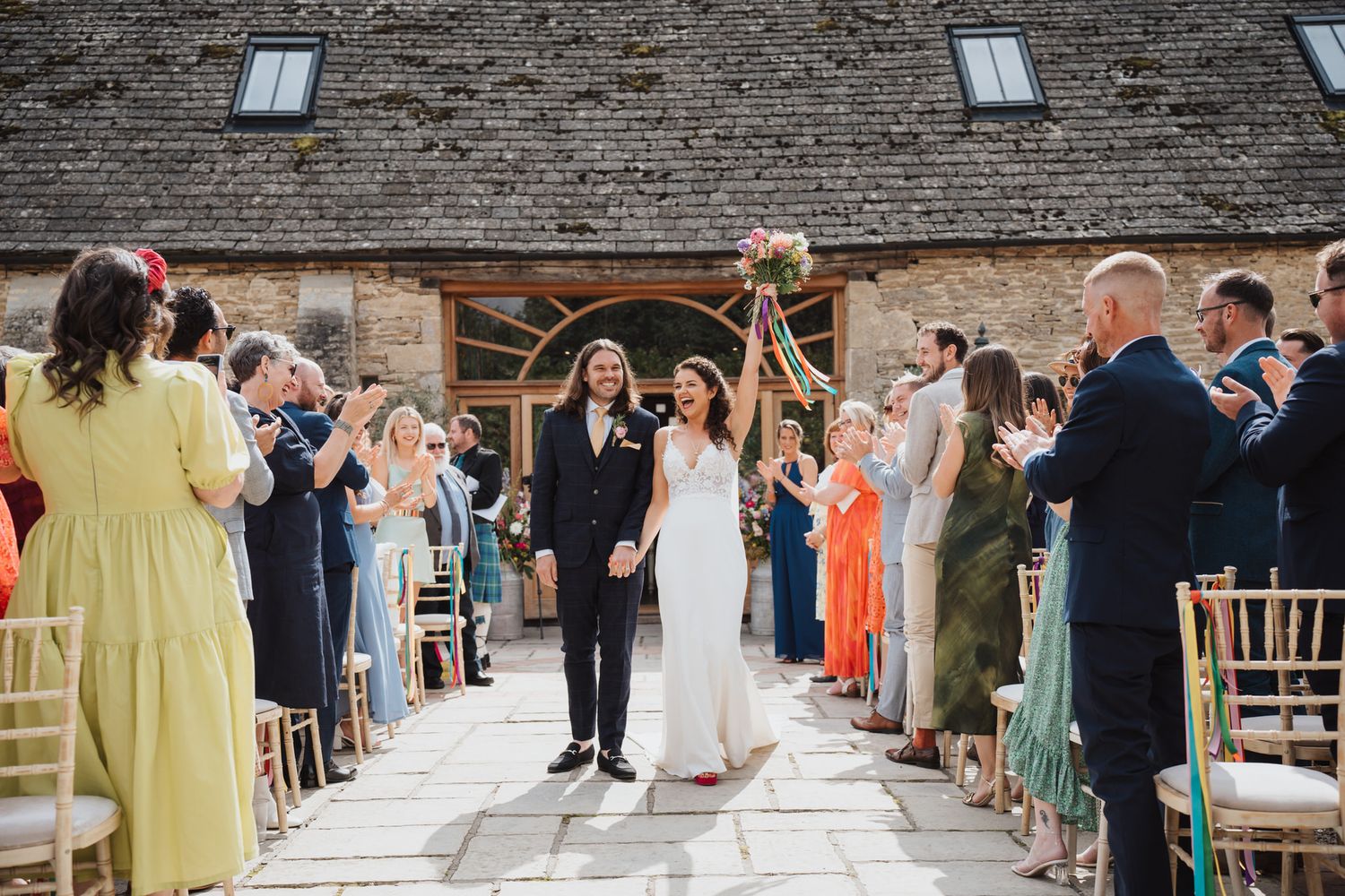 Bride and groom walk down the aisle at an outdoor barn wedding ceremony, greeted by cheering guests.