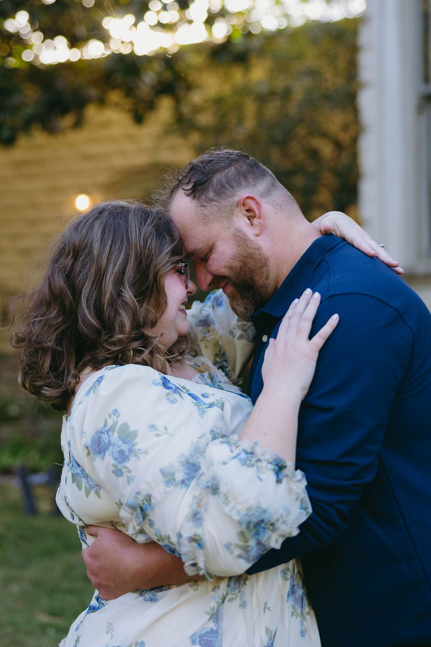 Couple embracing outdoors at dusk, woman in floral dress and man in navy suit touching foreheads romantically.