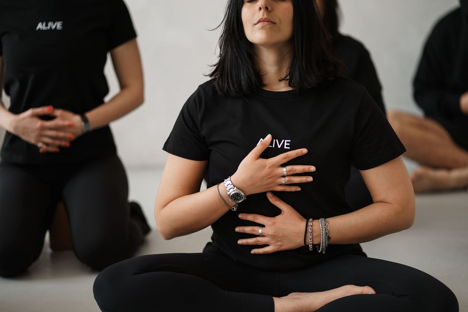 Group meditation session with people in black Alive t-shirts sitting cross-legged, hands on abdomen in mindful breathing pose.