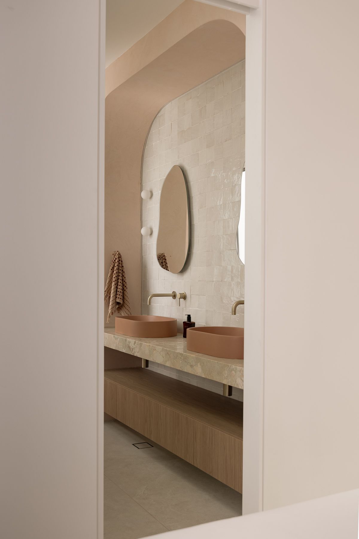 Modern bathroom with terracotta vessel sinks, arched mirror, brass faucets, and textured white tile wall in neutral tones.