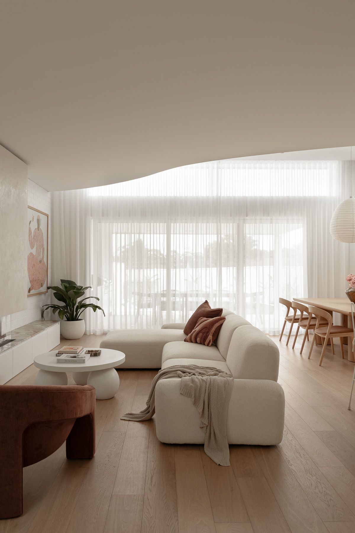 Modern minimalist living room with white curved sofa, sheer curtains, light wood floors, and neutral tones.
