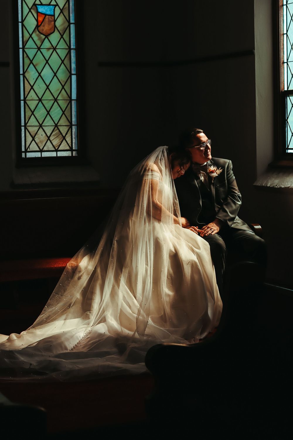 Bride and groom sitting together in a sunlit church pew, with stained glass windows casting light on her flowing veil.