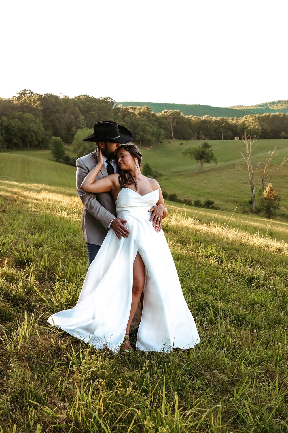 Bride in white gown with slit and groom in black cowboy hat embrace in a sunlit green countryside field.