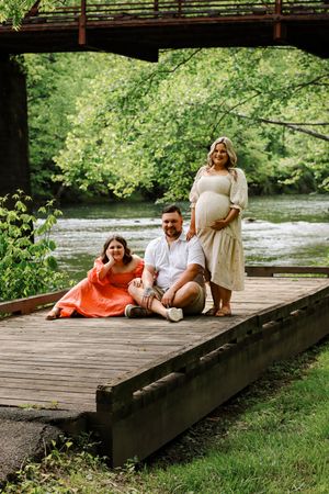 Pregnant woman in white dress poses with man and girl in orange dress on wooden deck by a river.