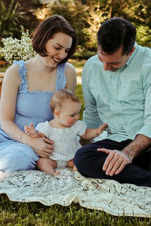 Happy young family sitting on lace blanket outdoors, parents smiling at baby in white outfit on sunny day.