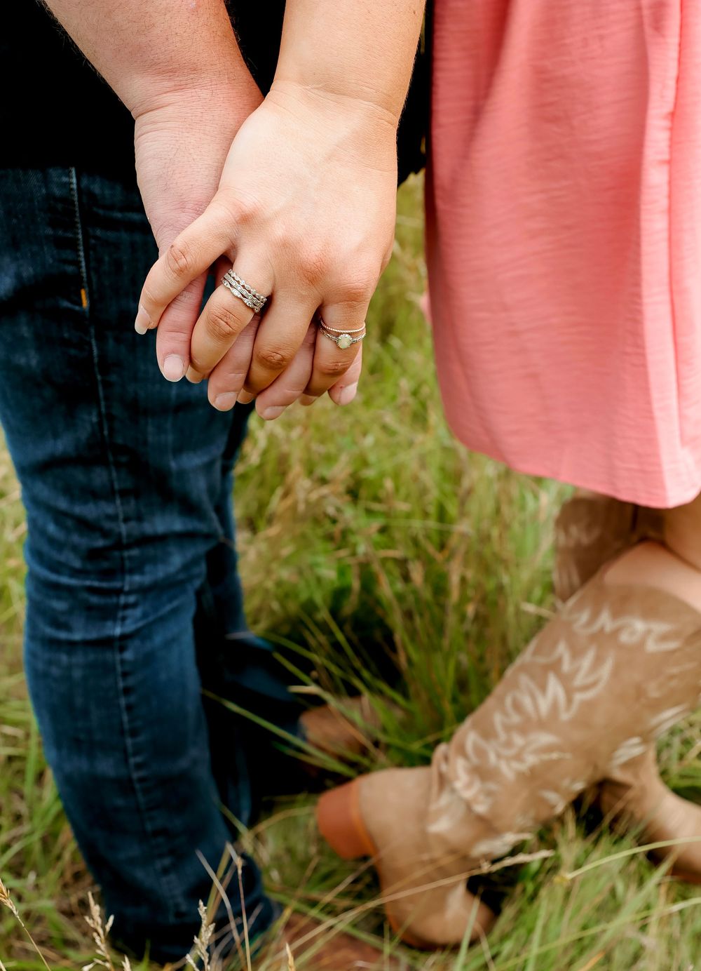 Couple holding hands with wedding rings, woman wearing cowboy boots and pink dress, standing in green grass field.