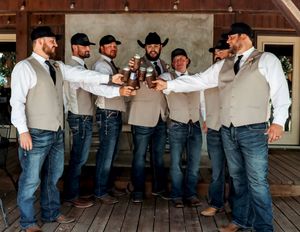 Seven groomsmen in cowboy hats, vests, and jeans toasting with beer bottles at a rustic outdoor wedding venue.