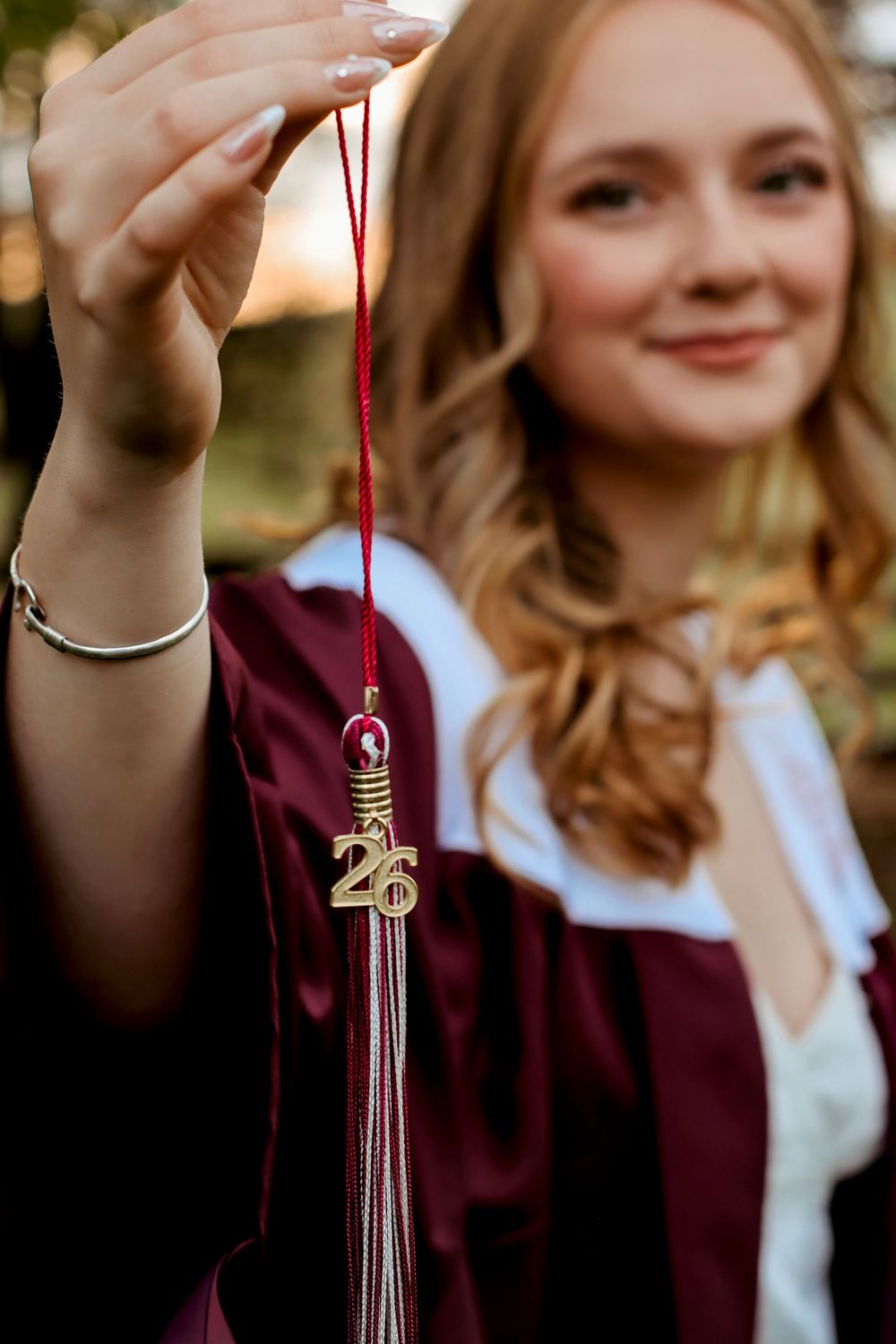 A smiling graduate in maroon cap and gown holds up a gold '26' tassel for her class of 2026 graduation photo.