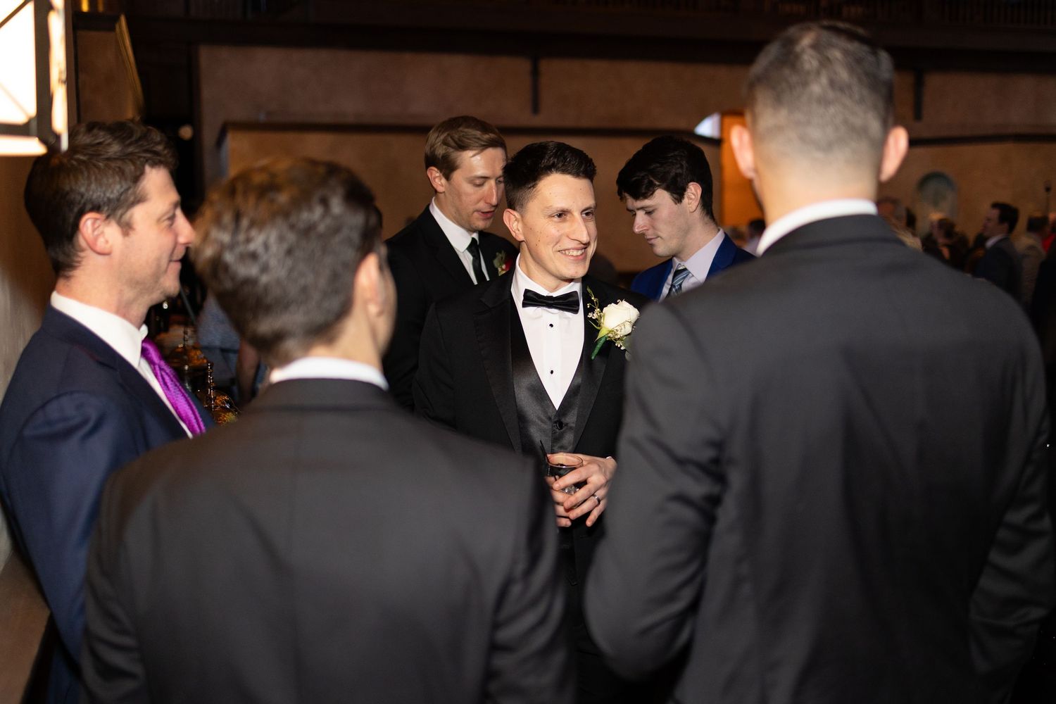 Groom in black tuxedo with boutonniere socializing with guests in suits at wedding reception in the Monastery event center in Cincinnati.