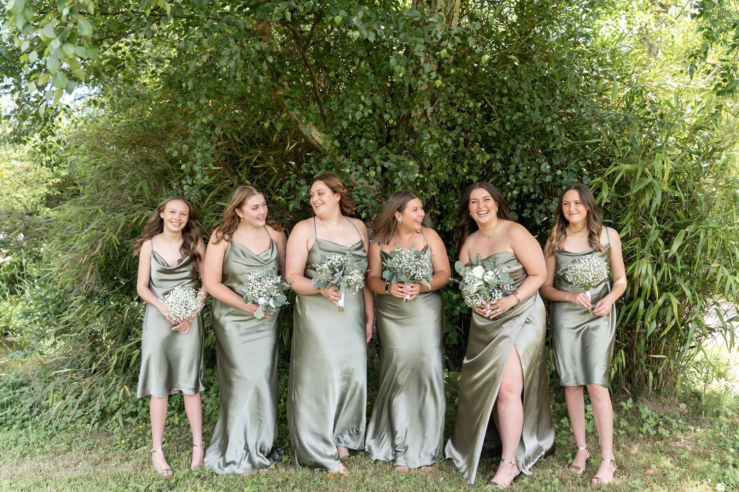 Six bridesmaids in sage green satin dresses holding greenery bouquets standing outdoors in front of lush green trees.