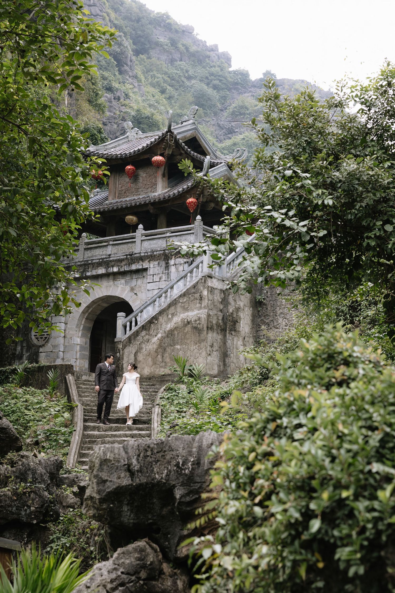 Couple in wedding attire poses on stone steps before a traditional Asian pagoda surrounded by lush greenery and red lanterns.