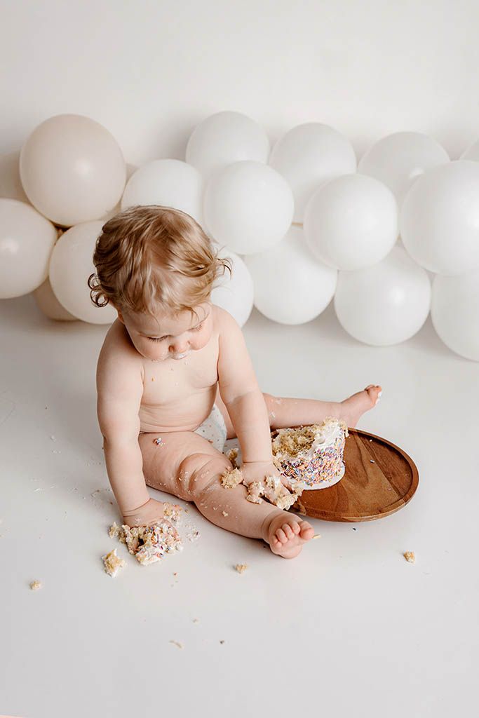 Baby smashing first birthday cake on brown plate surrounded by white balloons in a bright studio setting.