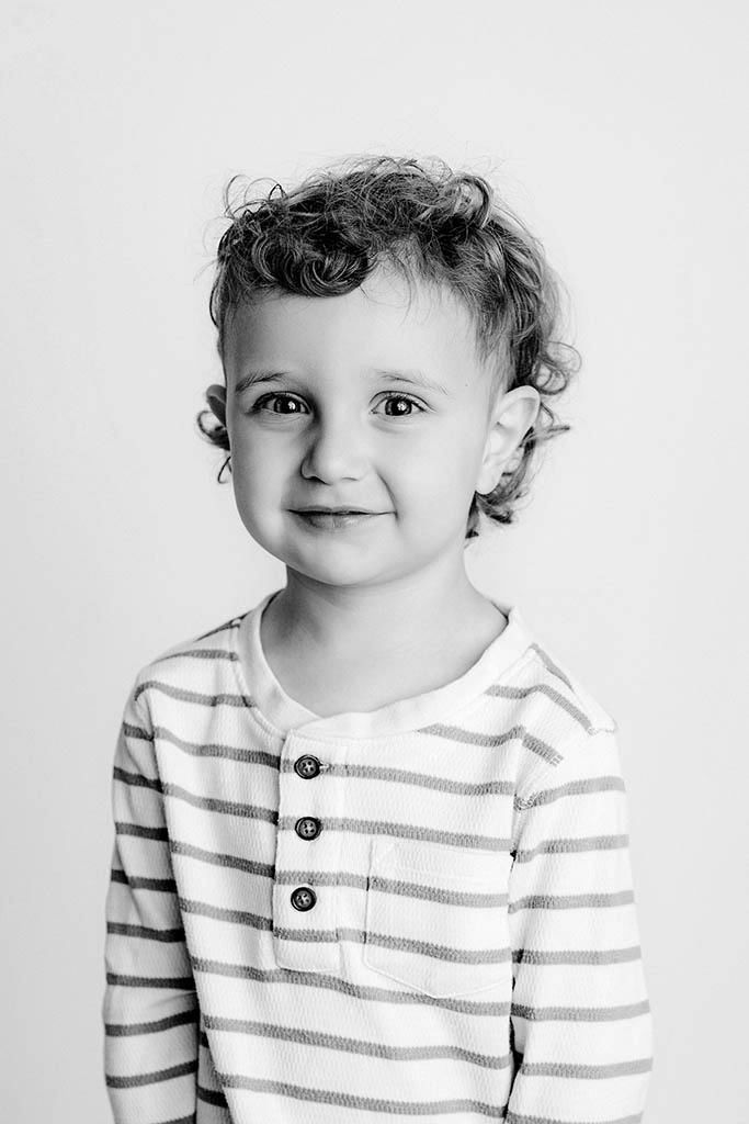Black and white portrait of a curly-haired toddler in a striped henley shirt smiling softly at the camera.