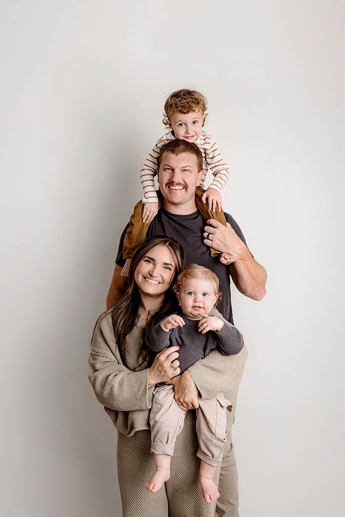 Family portrait of parents with two young children, one on dad's shoulders, against a white background.
