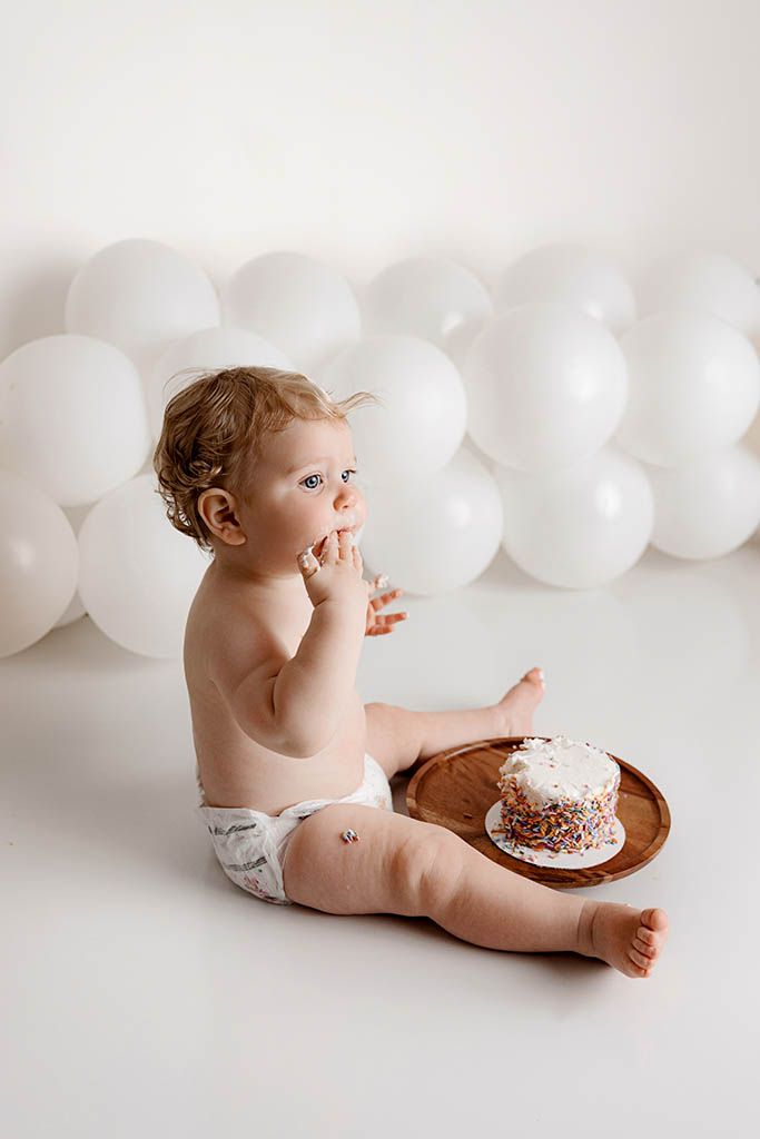 Baby in diaper eating smash cake on wooden plate, surrounded by white balloons, first birthday photo session.