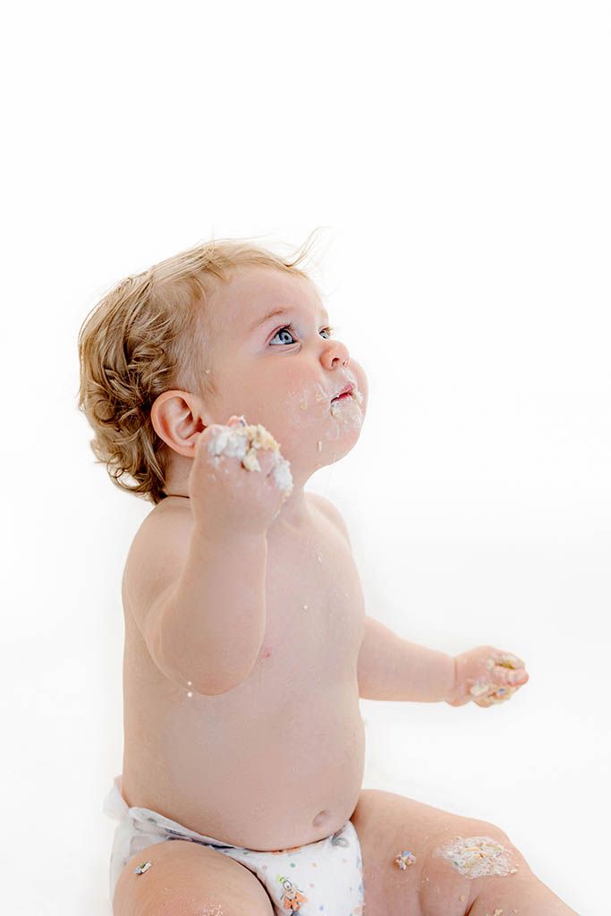 A messy baby in a diaper sits against white background, covered in cake frosting, looking up curiously.