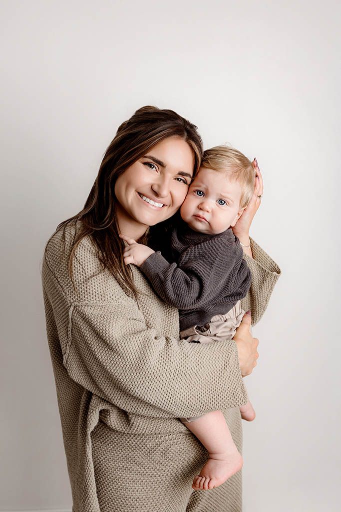 Smiling brunette woman in olive knit outfit holds a cute baby in a gray onesie against a white background.
