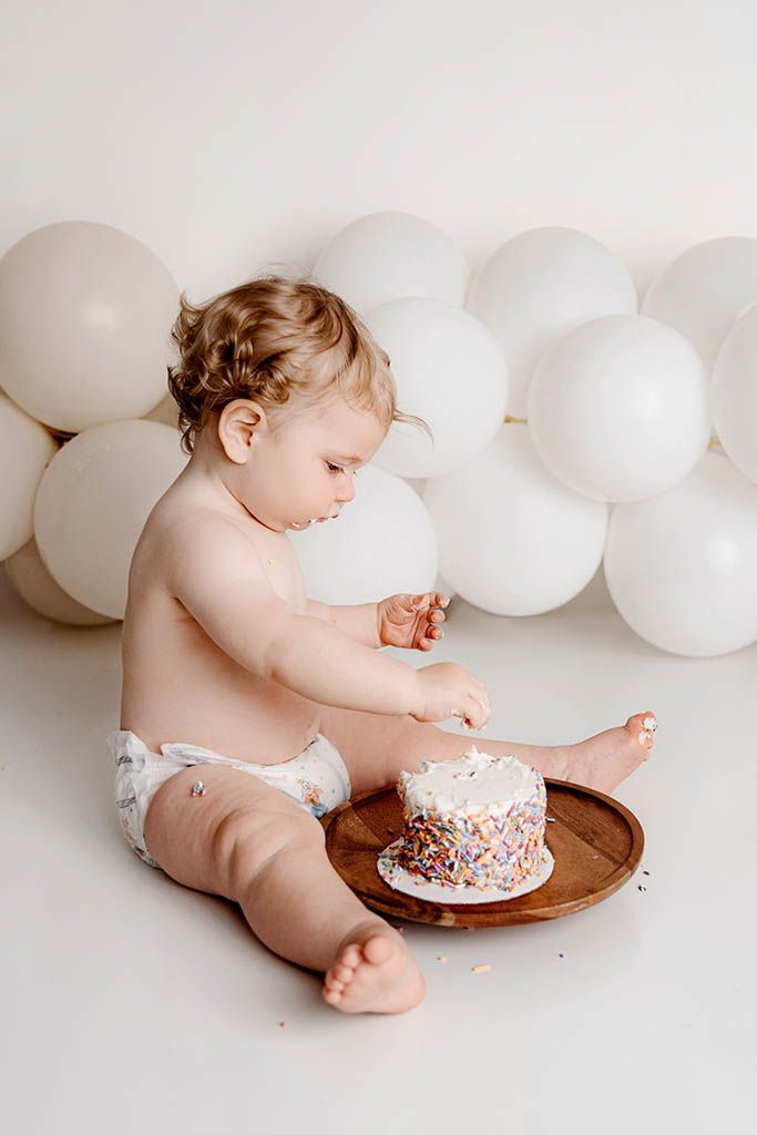 Baby in diaper touching sprinkle cake on wooden board, white balloons backdrop, first birthday cake smash photo.