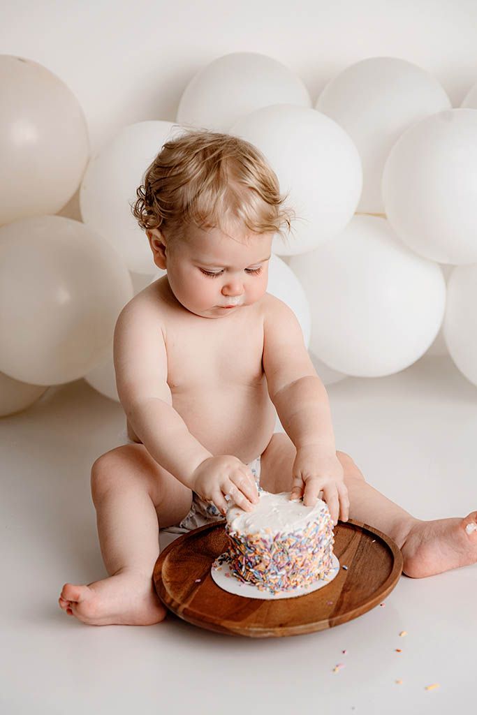 Baby smashing first birthday cake with white frosting and sprinkles, surrounded by white balloons on a wooden board.
