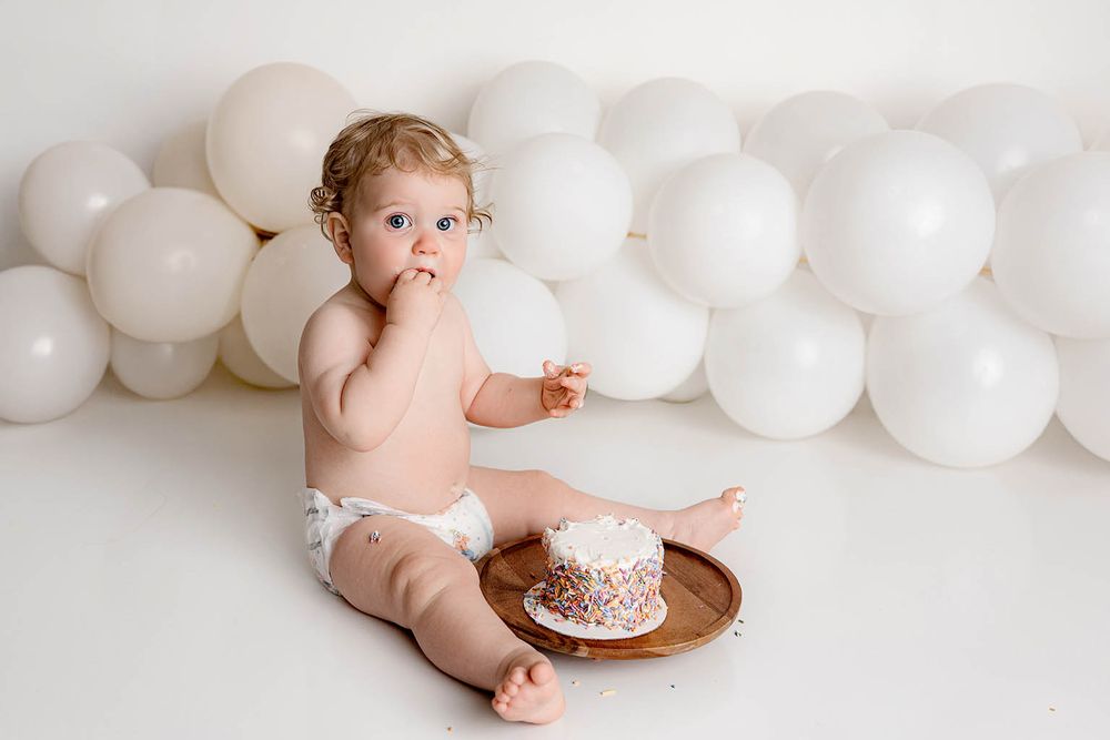 Baby in diaper sitting on floor with smash cake on wooden tray, surrounded by white balloons during first birthday celebration.
