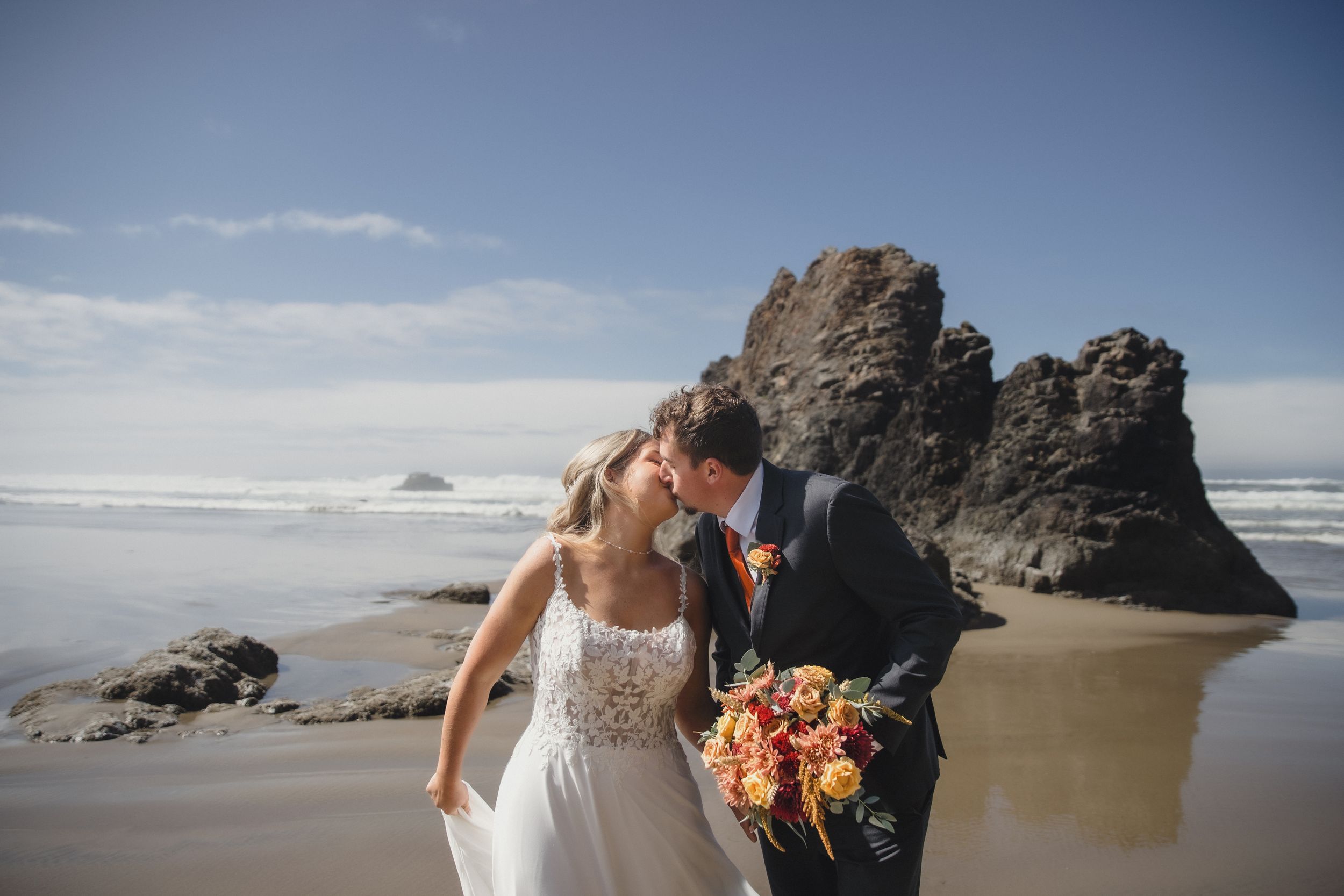 Bride and groom share a romantic kiss on a scenic beach with dramatic rock formations and ocean waves in the background.