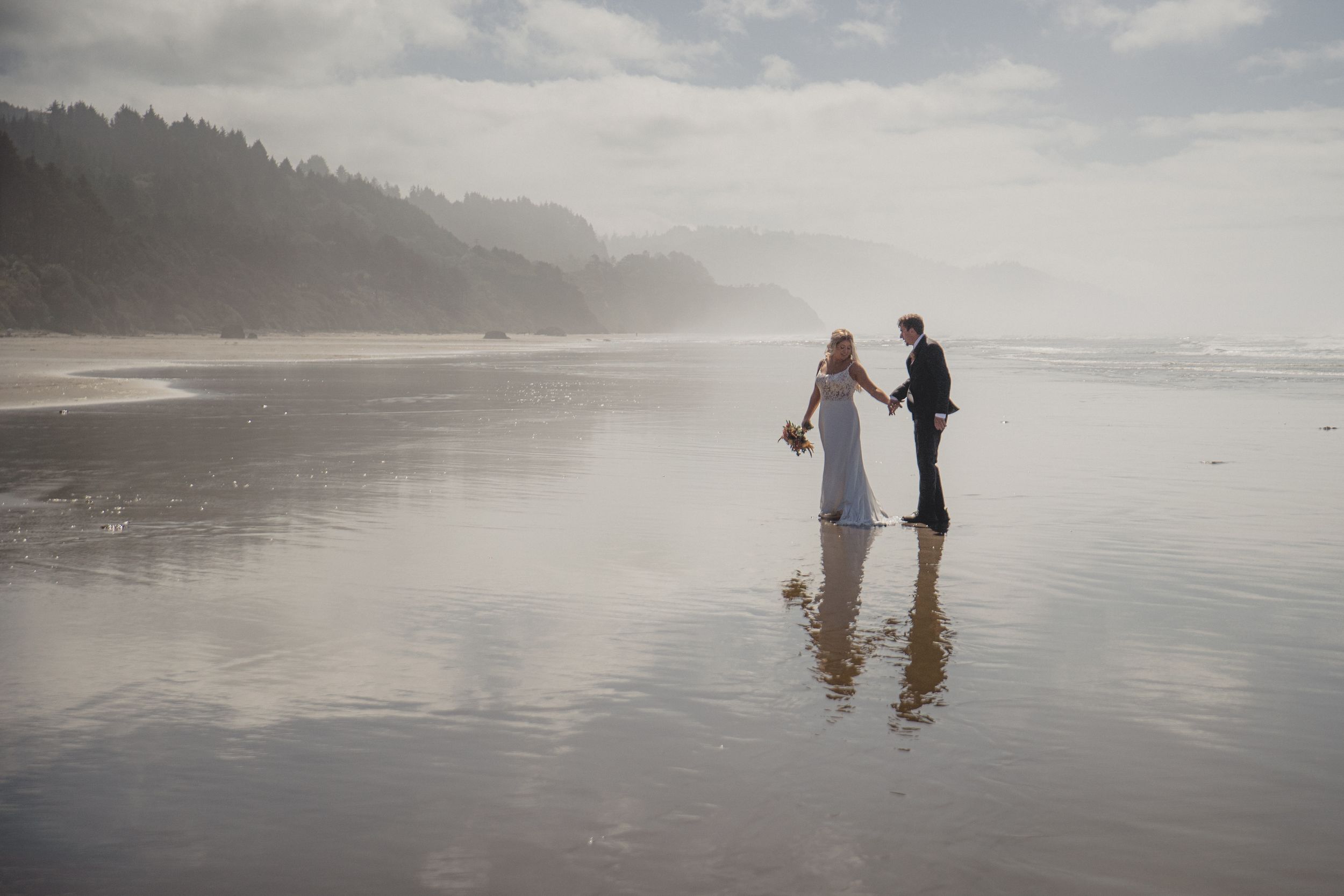 A bride and groom hold hands on a misty reflective beach with forested cliffs in the background.