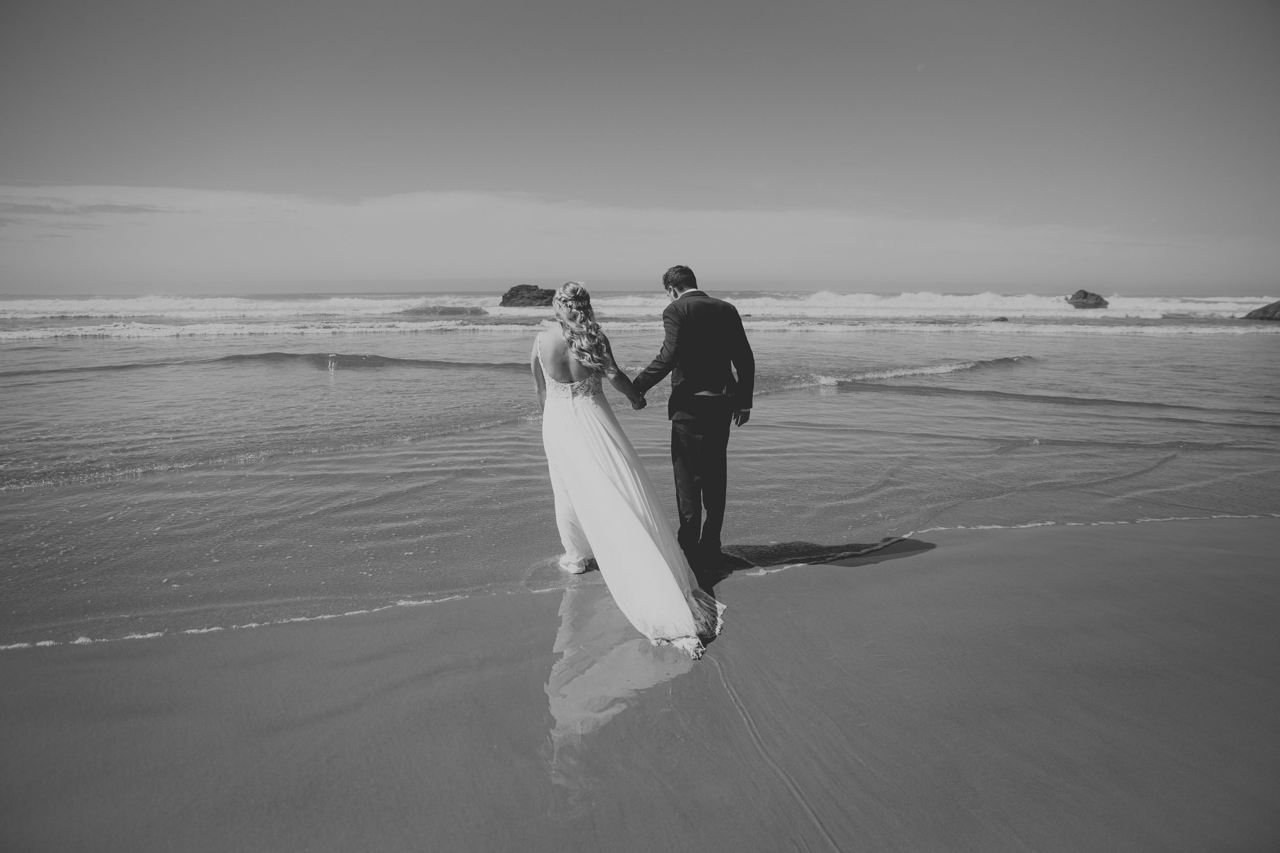 Black and white photo of a bride and groom holding hands walking along a sandy beach with ocean waves behind them.